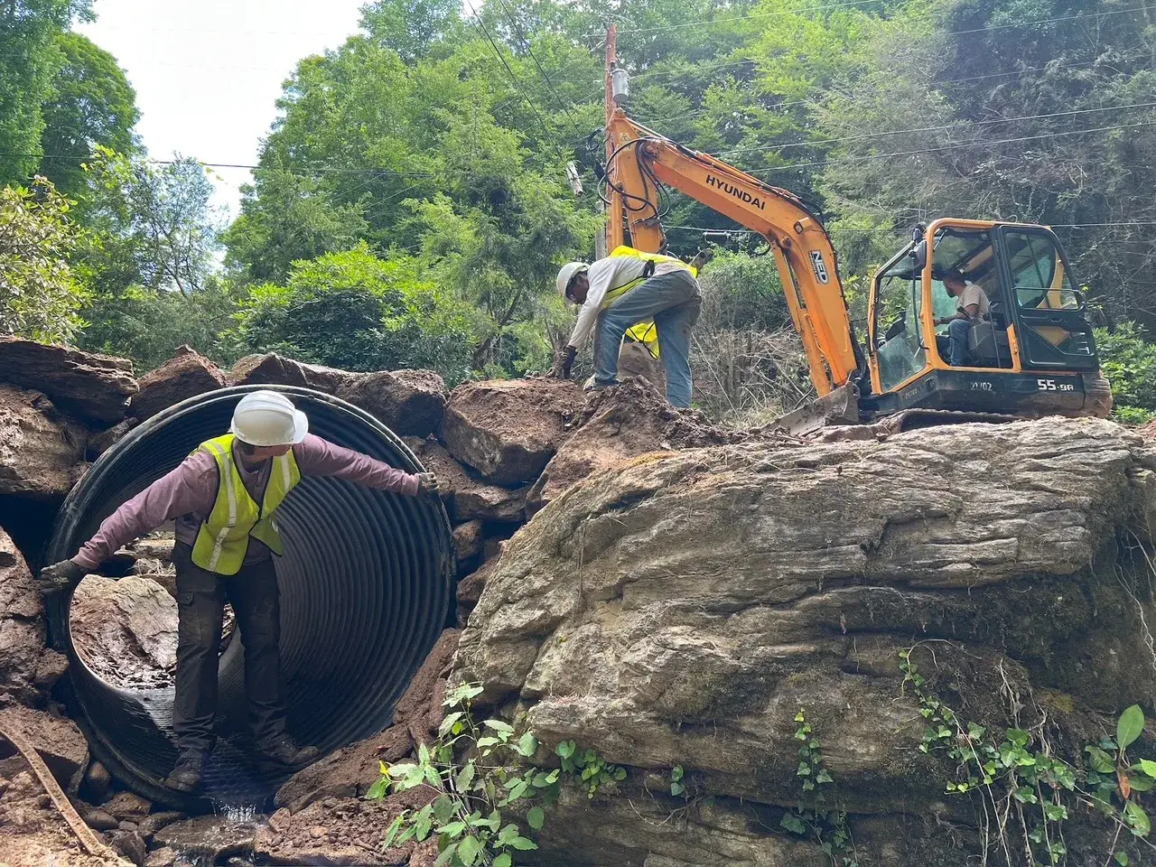 An excavator digging beside a large rock while a worker in a hard hat watches
